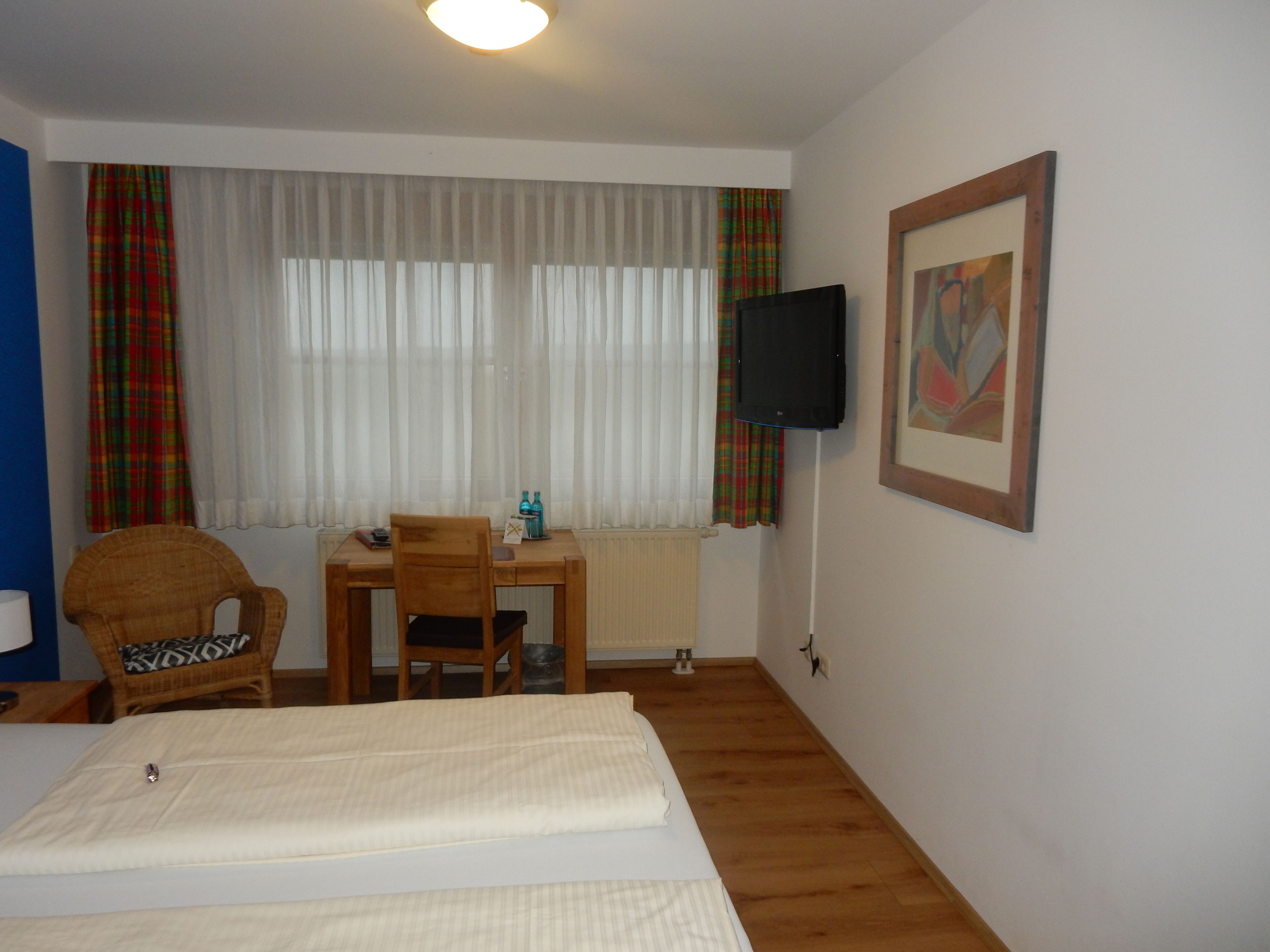 A hotel room with a bed, a desk with a chair, a television mounted on the wall, and a window with curtains.