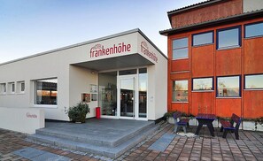 Un hotel llamado 'Hotel Frankenhöhe' con una fachada moderna, una entrada de cristal y una pequeña zona exterior con bancos.