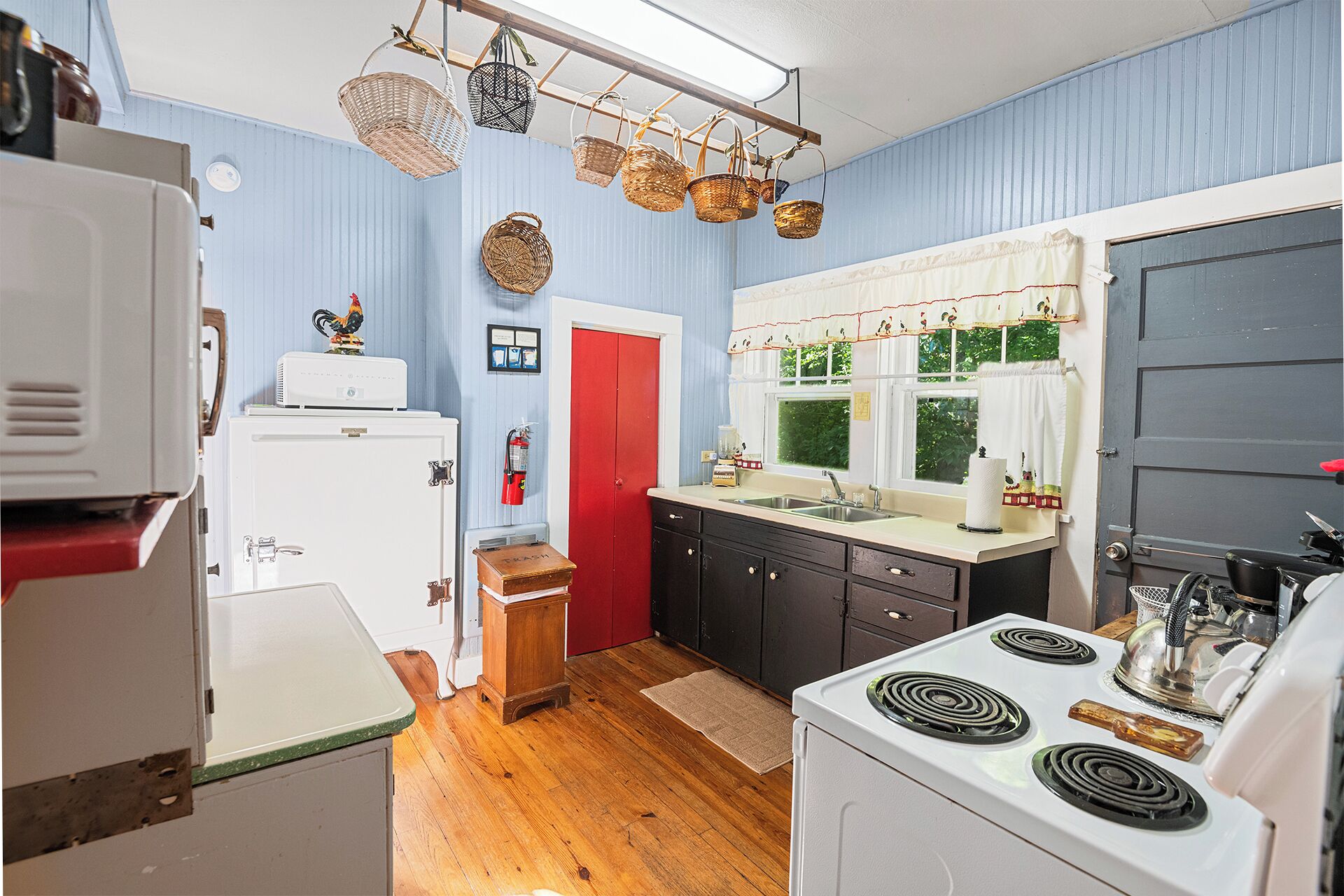 A kitchen with a stove, sink, and cabinets, a red door, and hanging baskets.