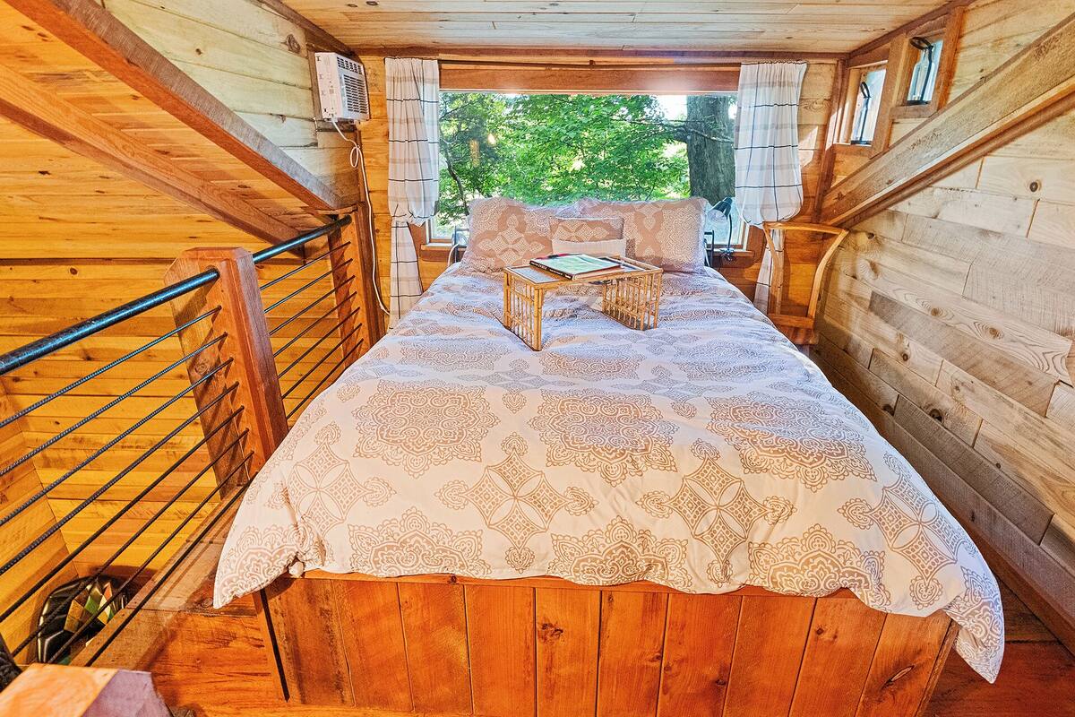 A wooden cabin interior with a bed, a small table, and a window with a view of trees.