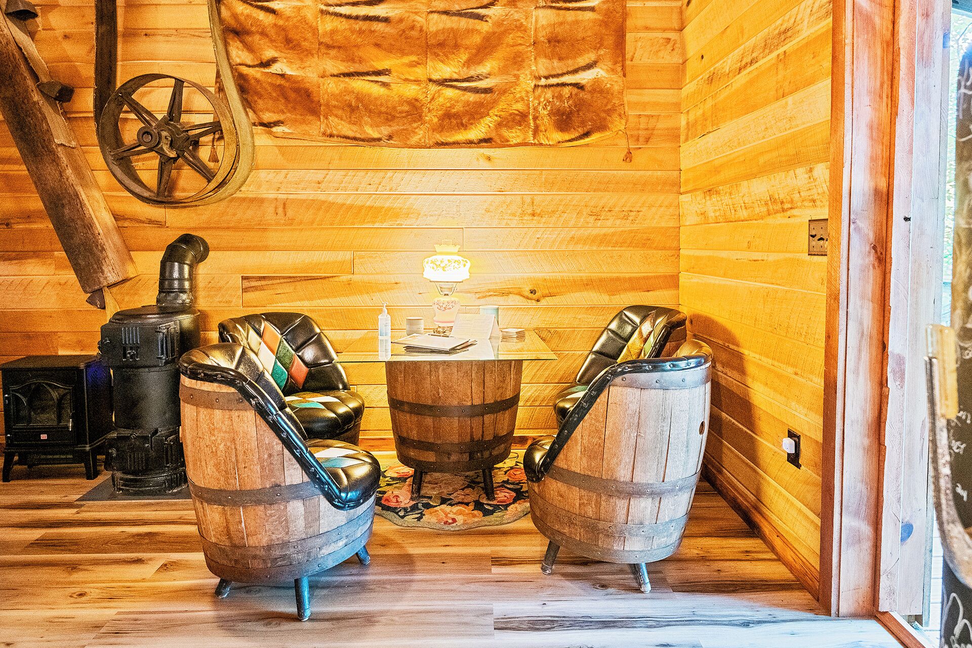 A wooden cabin interior with two leather chairs, a table, a fireplace, and a fur wall hanging.