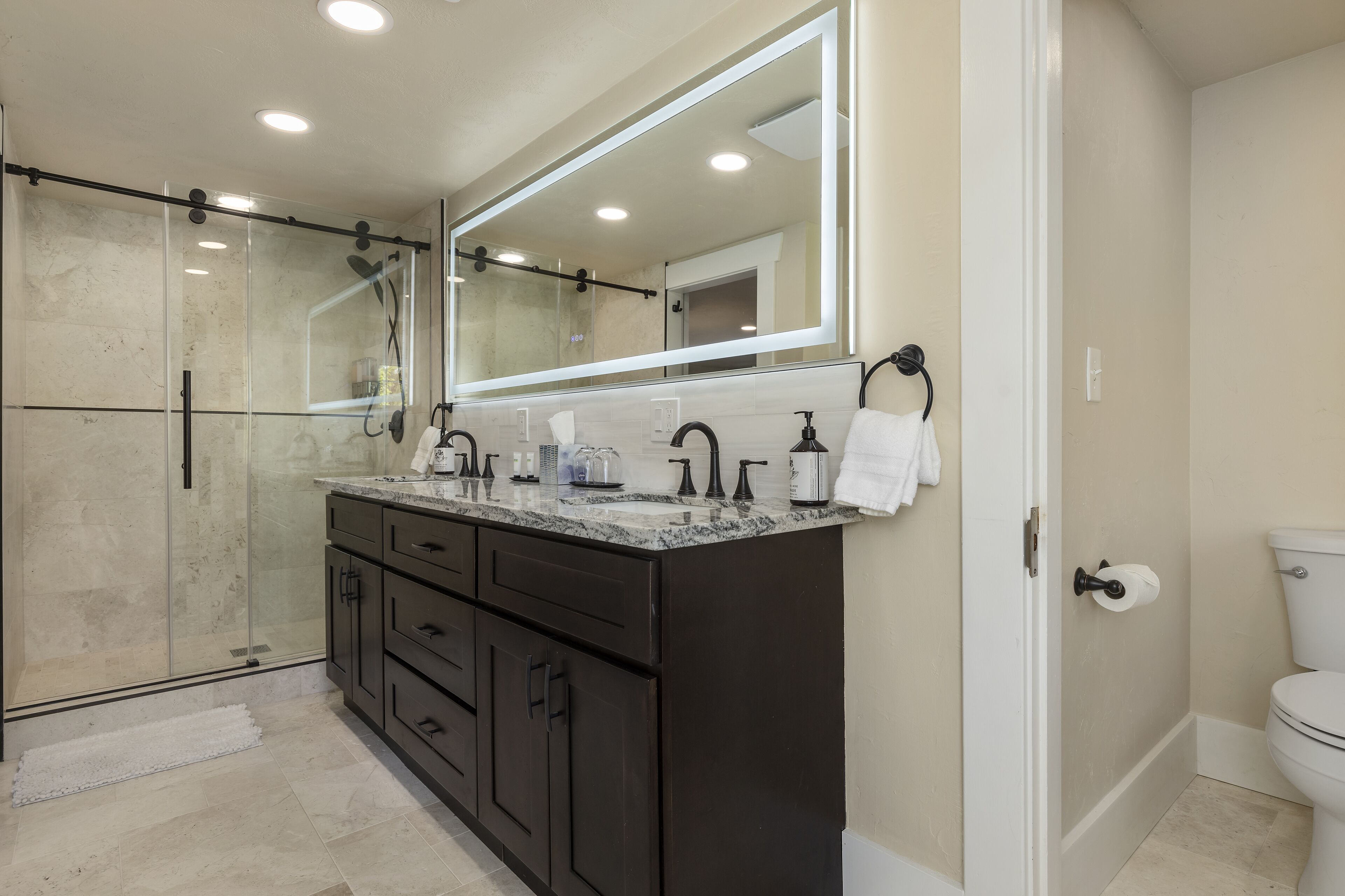 A modern bathroom with a glass-enclosed shower, dark cabinetry, a large mirror, and a toilet.