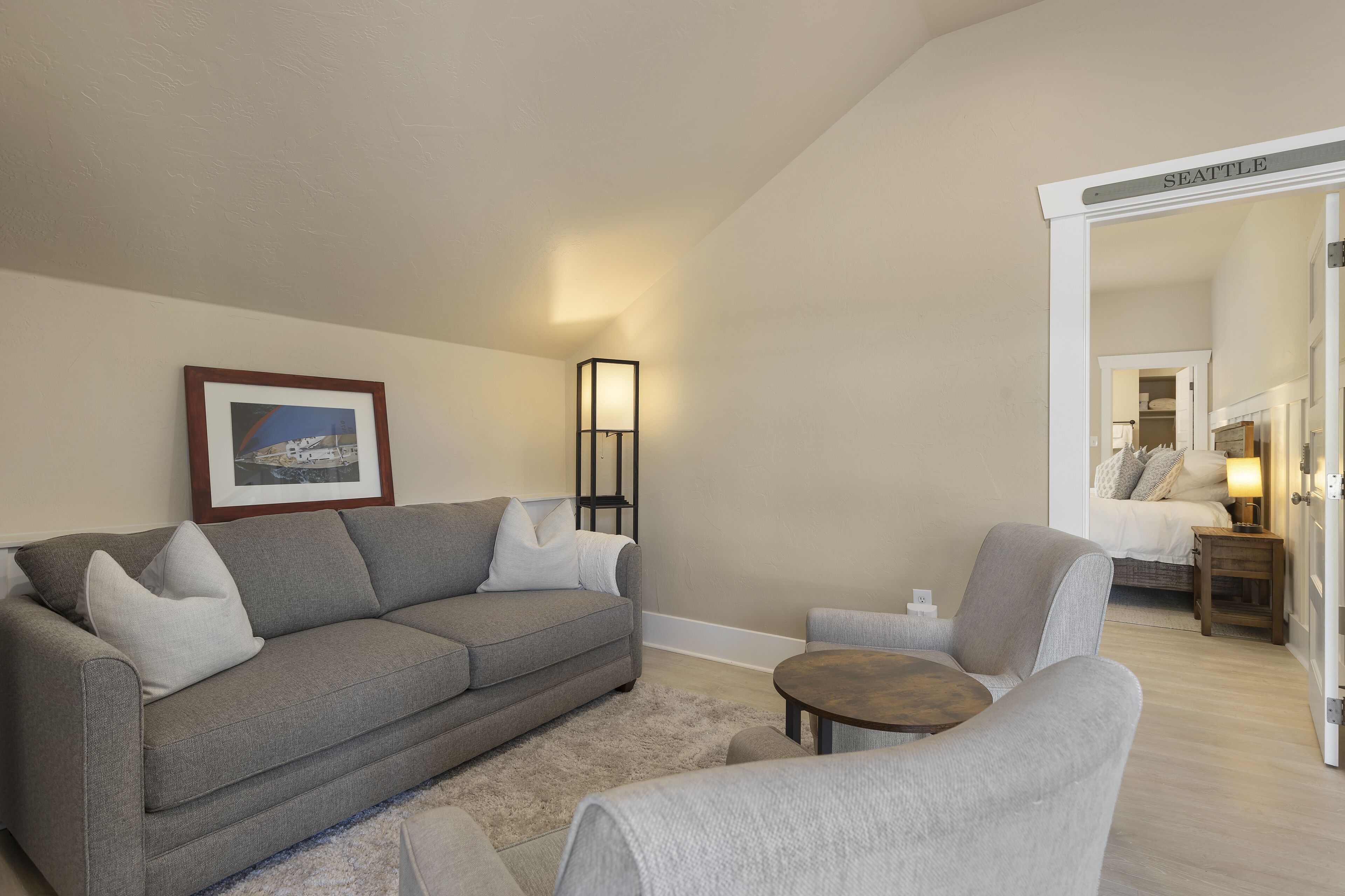 A living room with a grey sofa, a small round table, and a chair. There is a framed picture on the wall and a floor lamp.