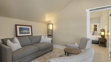 A living room with a grey sofa, a small round table, and a chair. There is a framed picture on the wall and a floor lamp.
