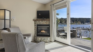 A living room with a stone fireplace, a flat-screen TV, and a view of a lake and dock. - Waterfront Inn (Gig Harbor)