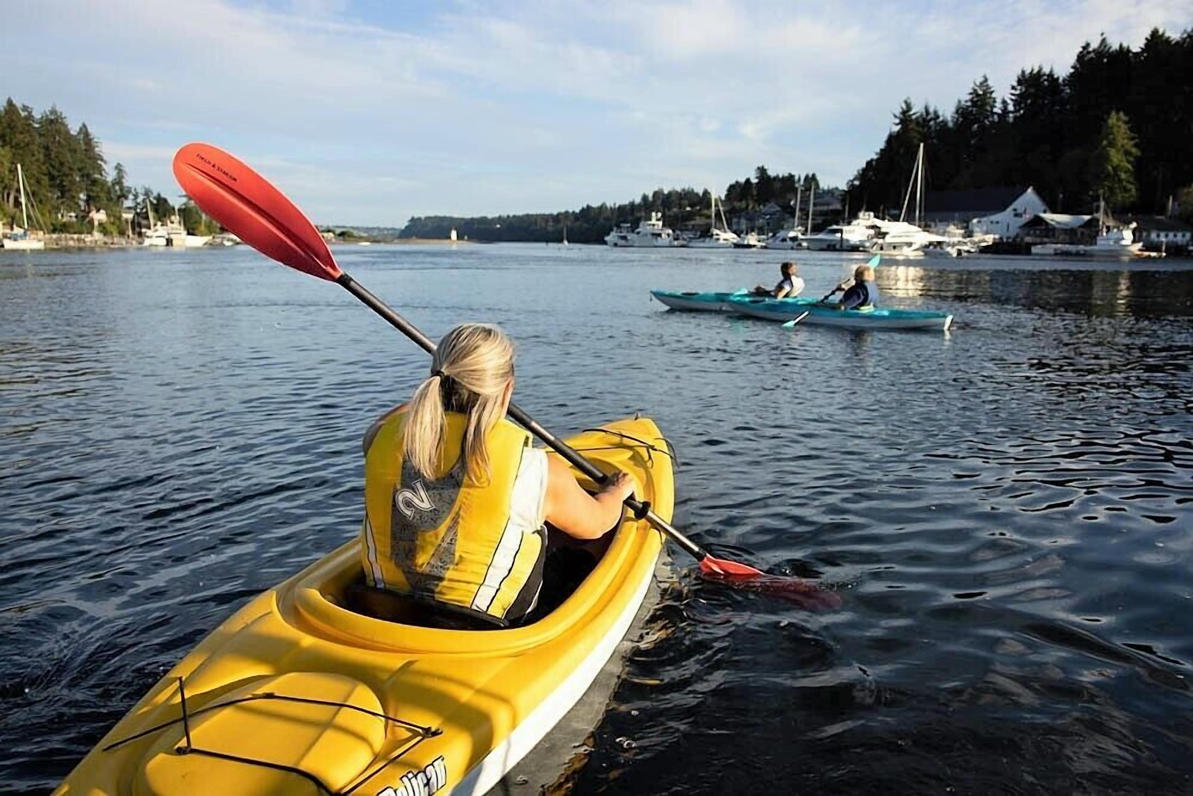 Una persona haciendo kayak en un lago tranquilo, con barcos y casas al fondo.