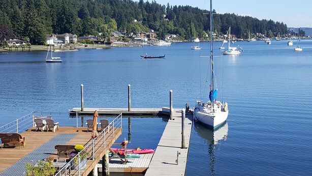 A marina with boats docked, a wooden pier with seating, and a backdrop of houses and trees. - Waterfront Inn (Gig Harbor)