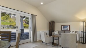 A modern living room with a sofa, armchairs, a coffee table, and a dining table. There is a sliding glass door leading to a balcony with a stone wall and greenery outside. - Waterfront Inn (Gig Harbor)