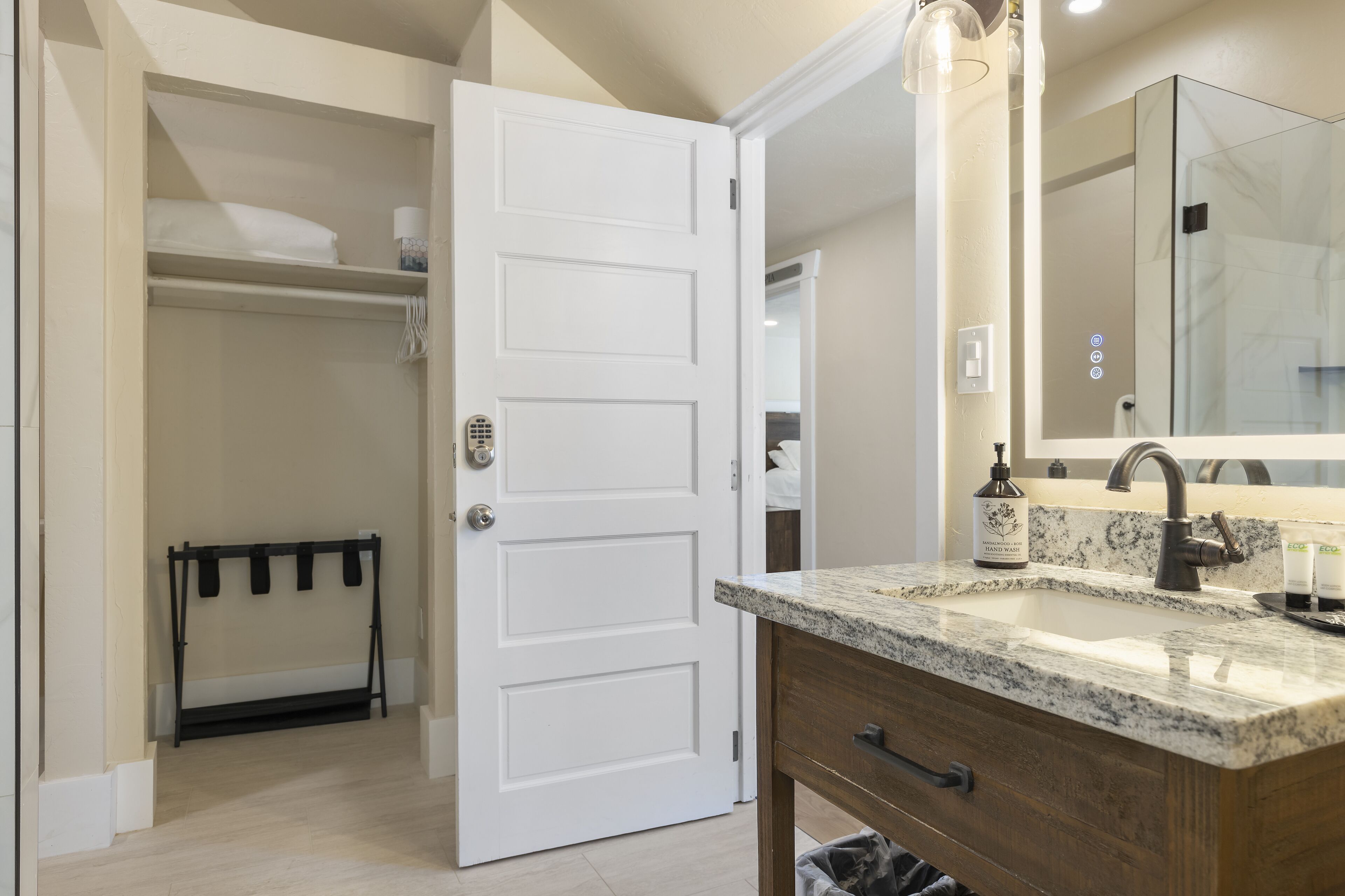 A bathroom with a white door, a vanity with a granite countertop, a mirror, and a shower area visible through the door.