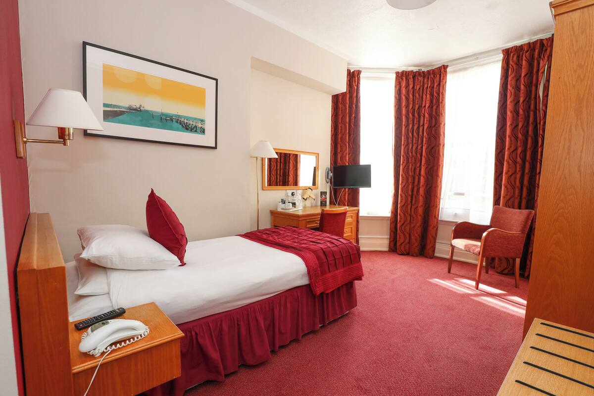 Een hotelkamer met een bed, een bureau met stoel, een televisie, een telefoon en een raam met gordijnen.