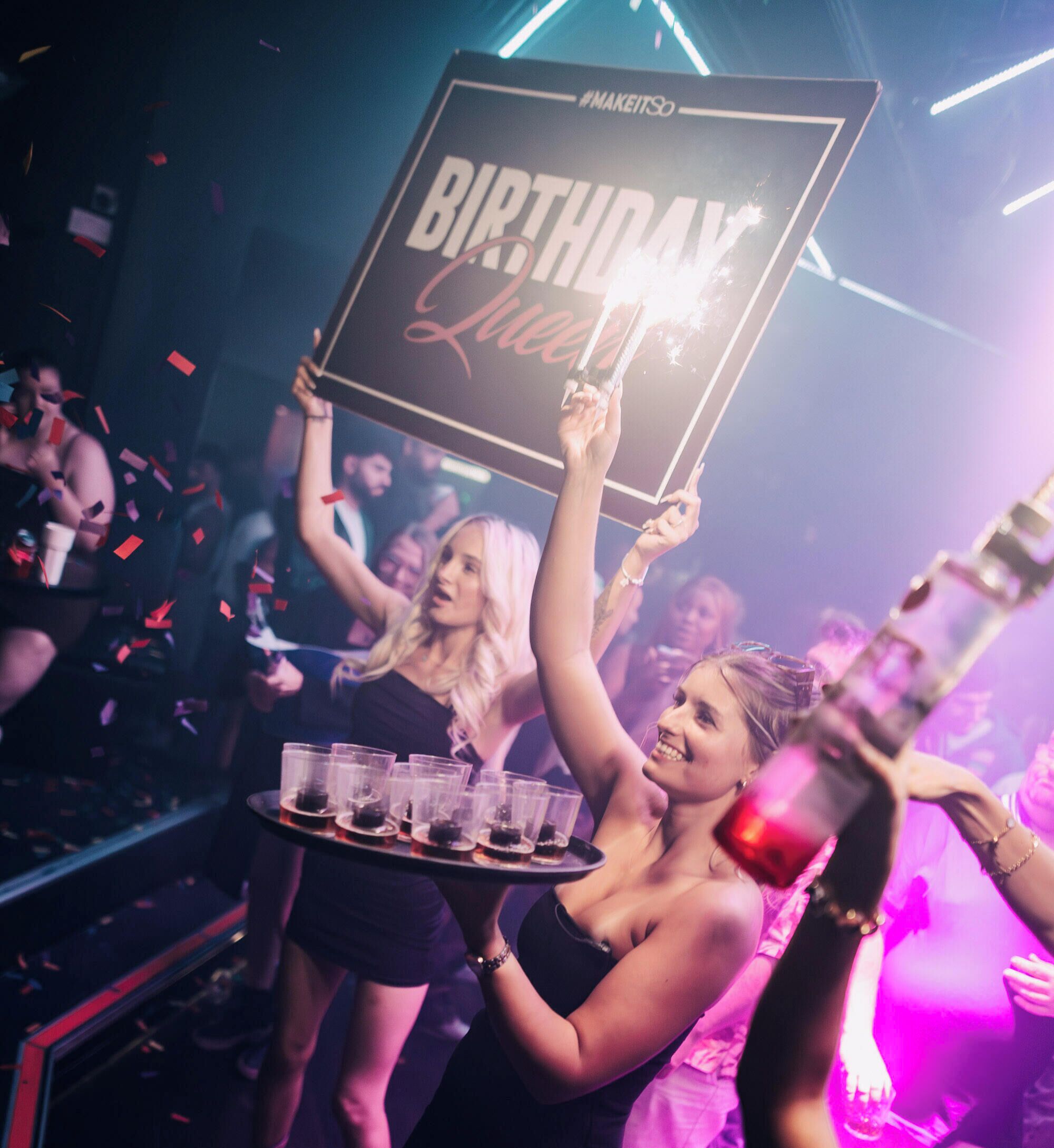 A person holding a sign that reads 'BIRTHDAY' with a party atmosphere in the background.