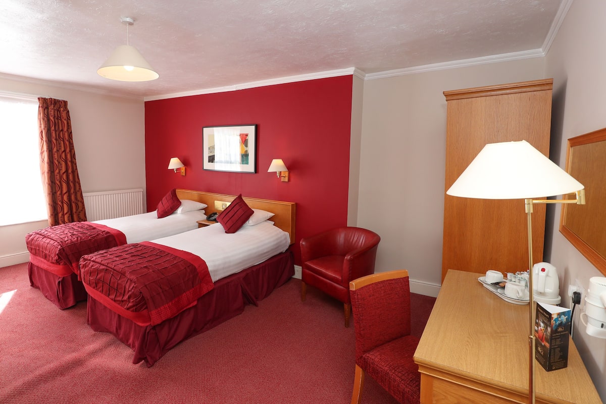 Hotelkamer met twee bedden, een rood tapijt, een houten garderobe, een bureau met lamp en een rode stoel.