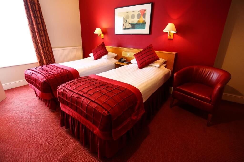 A hotel room with two beds, red bedding, a red chair, and a painting on the wall.