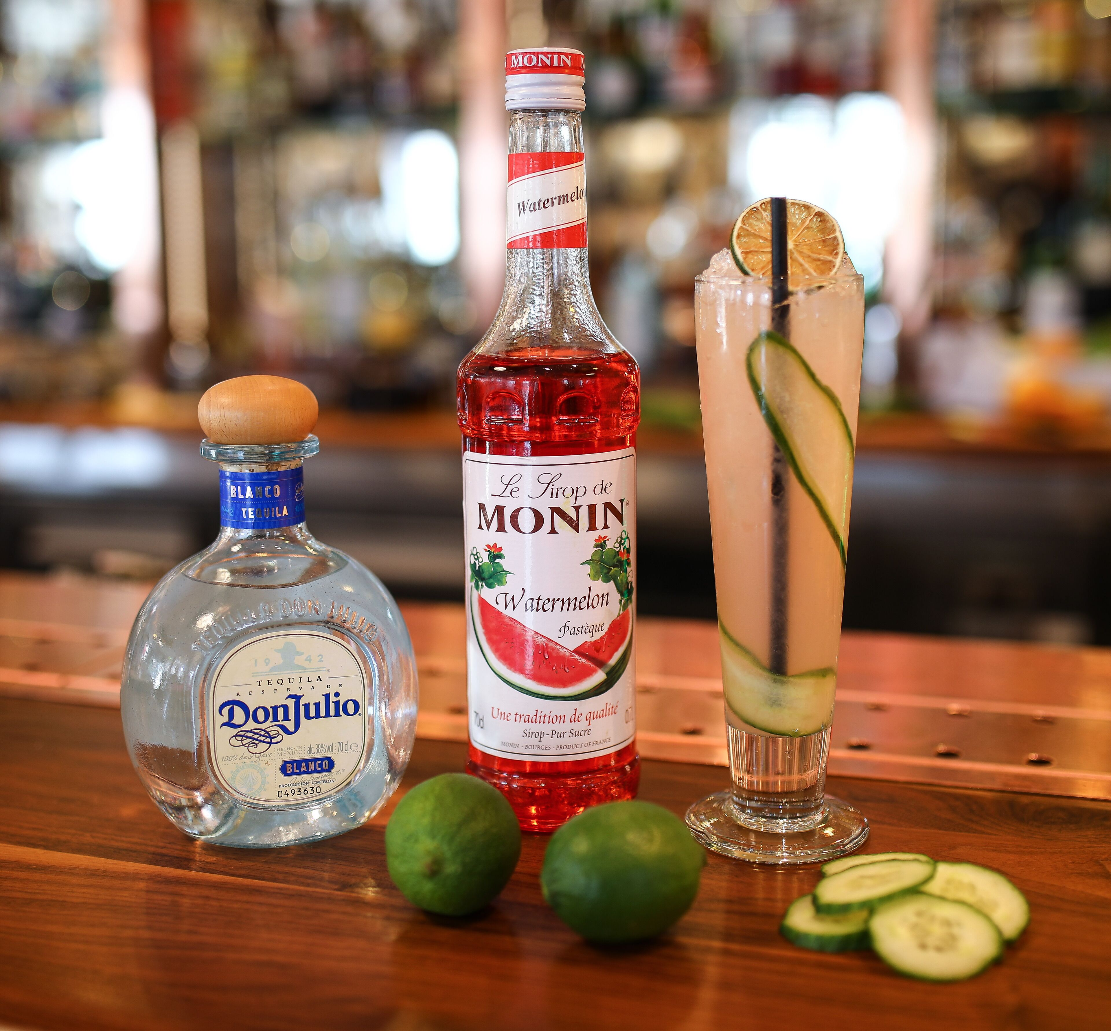 A bar setting with a bottle of Don Julio tequila, a bottle of Monin watermelon syrup, and a tall glass with a lime slice and cucumber garnish.
