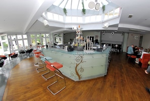 Ein großzügiger, moderner Barbereich mit hohen Decken und großen Fenstern, der eine zentrale Bartheke mit Hockern und ein dekoratives Baumdesign bietet.