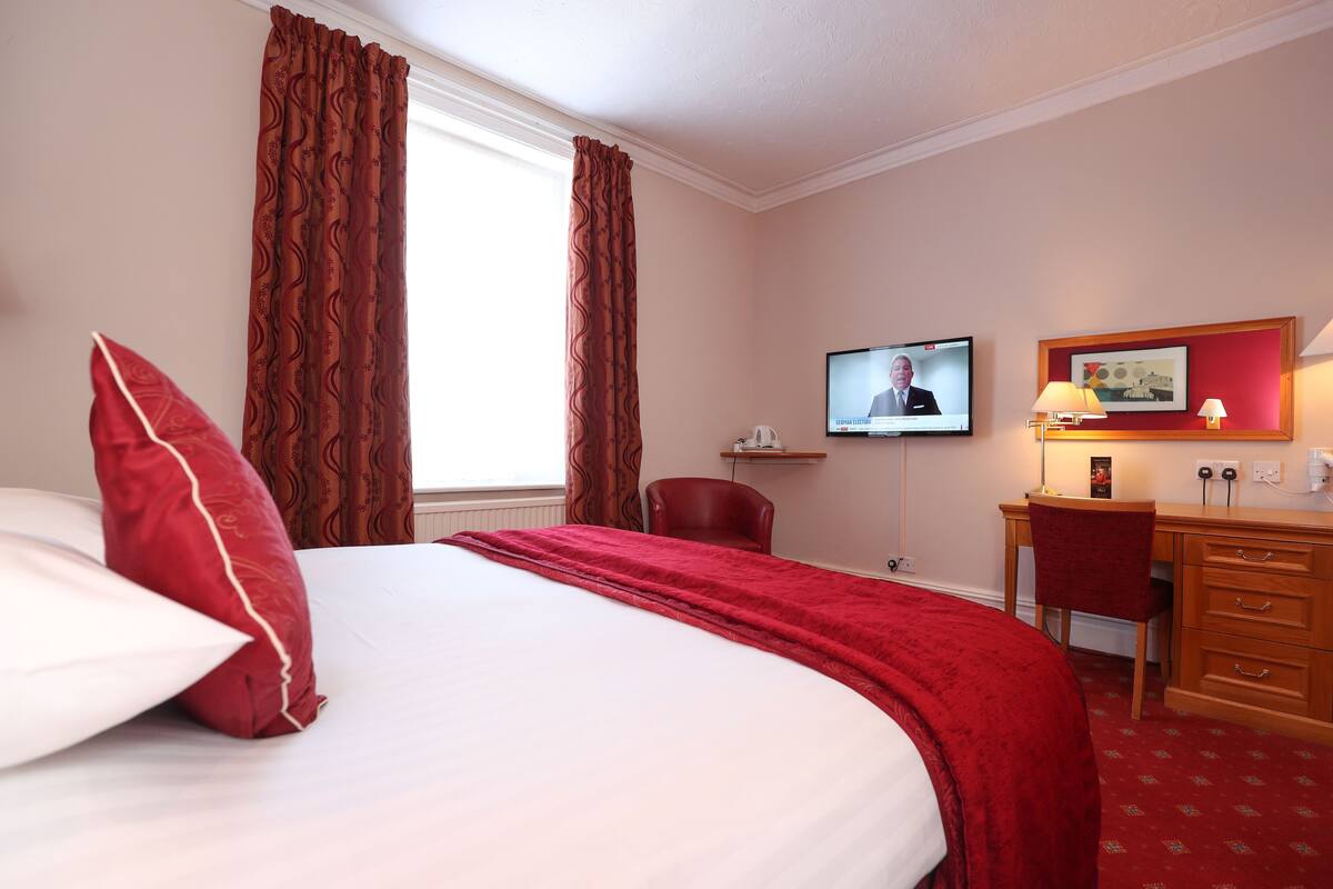 Een hotelkamer met een groot bed, een bureau met stoel, een televisie en een rode vloerbedekking.