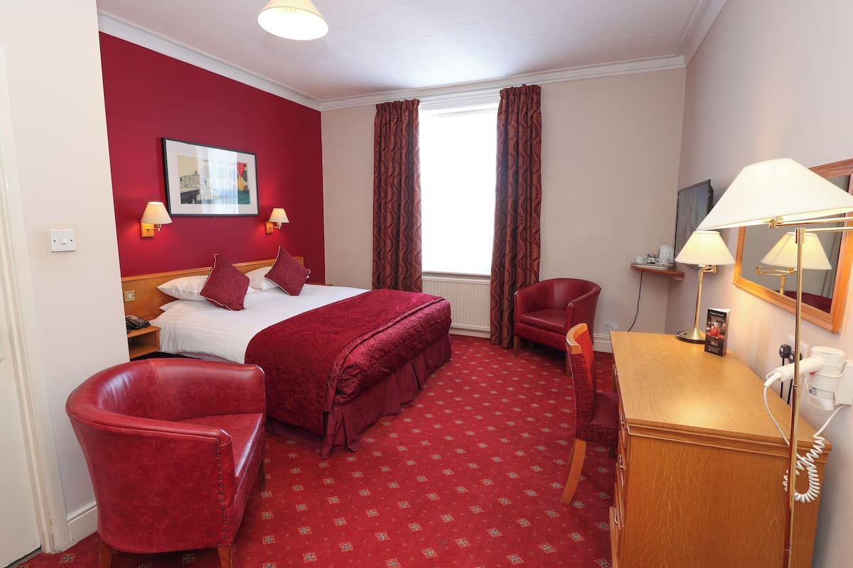 Een hotelkamer met een groot bed, twee rode stoelen, een bureau met lamp en een raam met gordijnen.