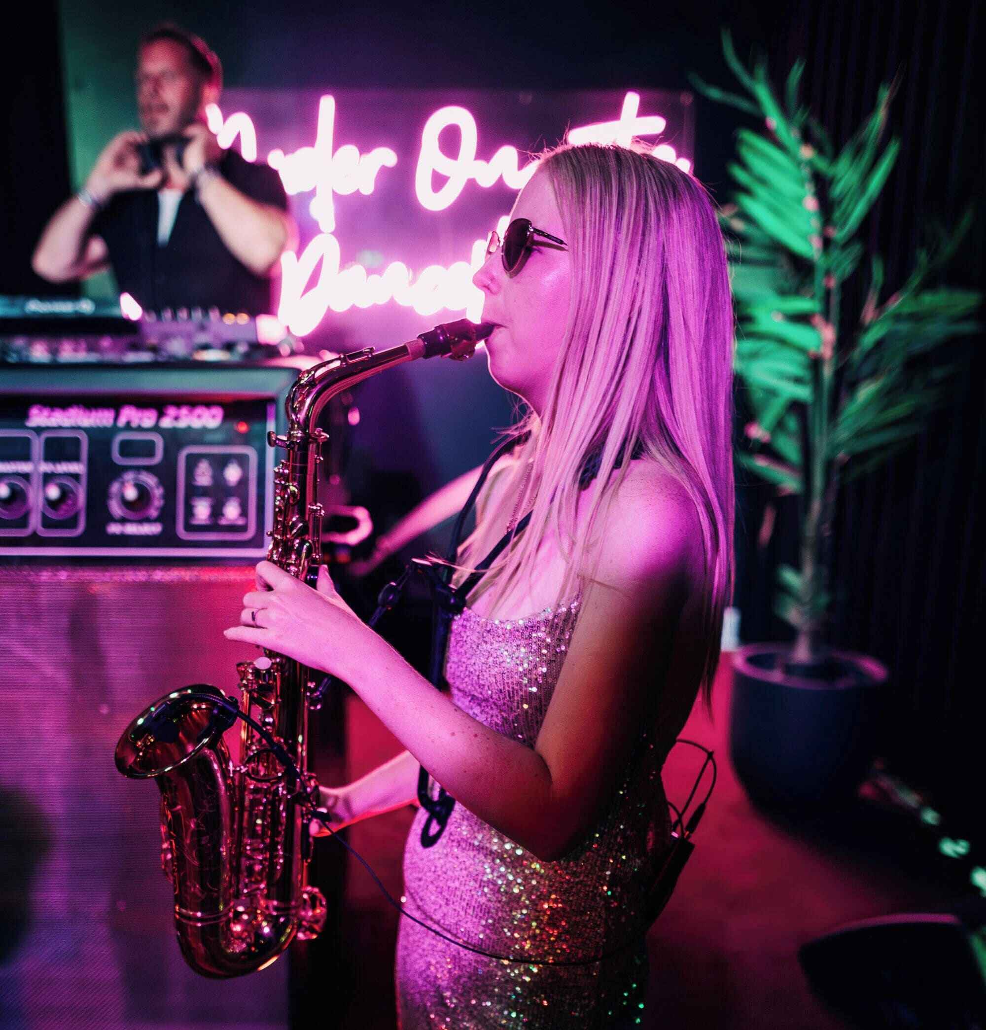 A person playing a saxophone in a room with a neon sign in the background.
