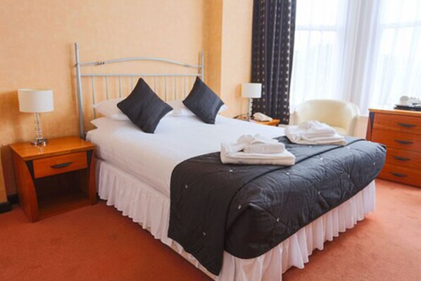 A hotel room with a bed, two bedside tables, a chair, a dresser, and a window with curtains. - Royal Exeter Hotel (Bournemouth)