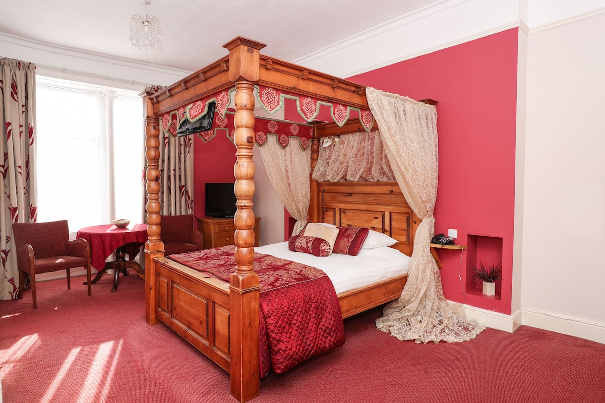 Een hemelbed met een rood-witte spreideken, een houten hoofdbord en een vloer met een rood tapijt.