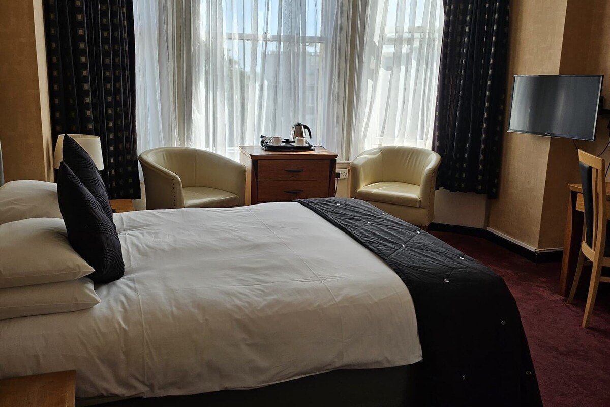 A hotel room with a bed, two chairs, a desk, a TV, and large windows with curtains.