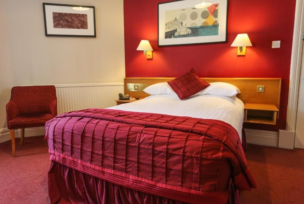 Una habitación de hotel con una cama, una silla, dos lámparas fijadas a la pared y un cuadro enmarcado.