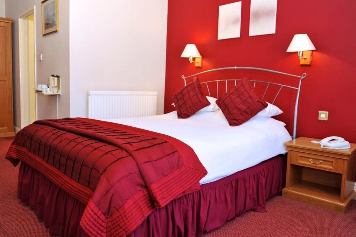 A hotel room with a bed, red walls, two bedside lamps, a wooden nightstand, and a radiator.