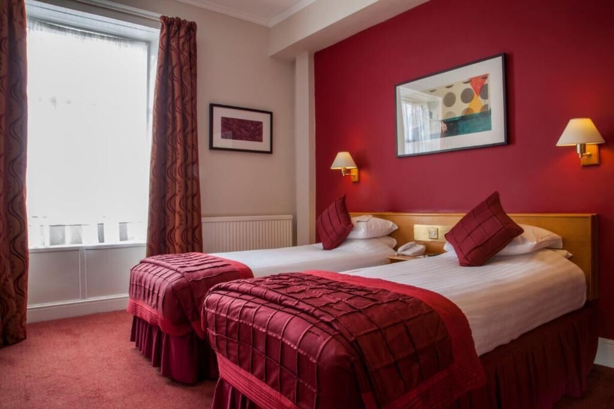 A hotel room with two beds, red curtains, a framed picture, and wall-mounted lamps.