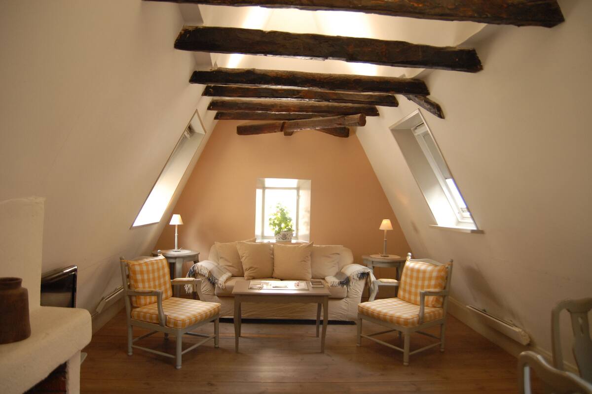 An attic room with a sloped ceiling, a sofa, two armchairs, a table, and a window.