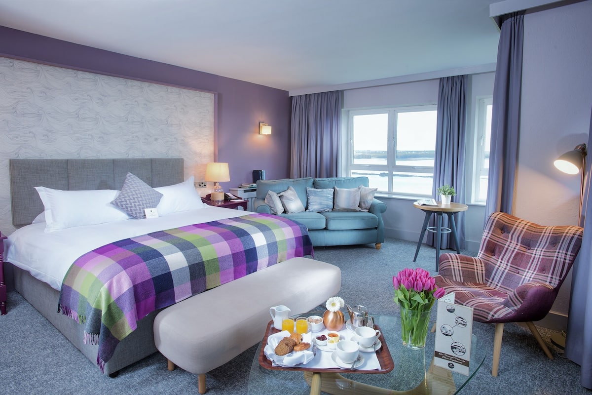 A hotel room with a bed, a sofa, a chair, a breakfast tray, and a window with curtains.