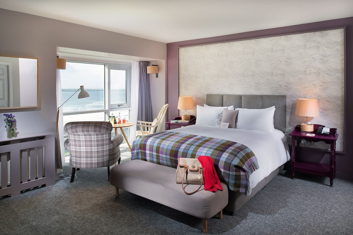 A hotel room with a large bed, a chair, a small table, and a view of the ocean.