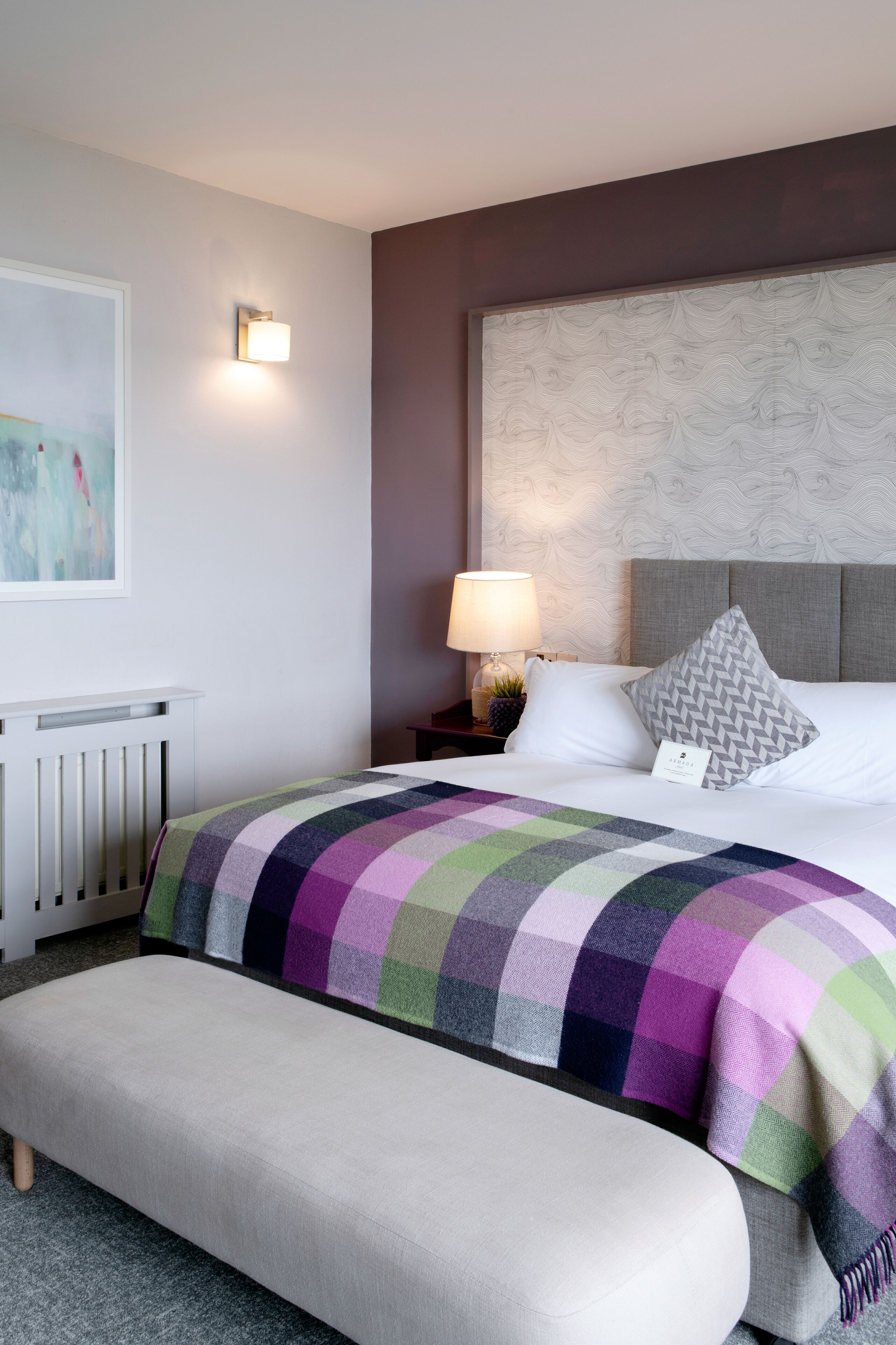 Une chambre d’hôtel comprenant un lit, une table de chevet avec une lampe, un banc et un luminaire fixé au mur.