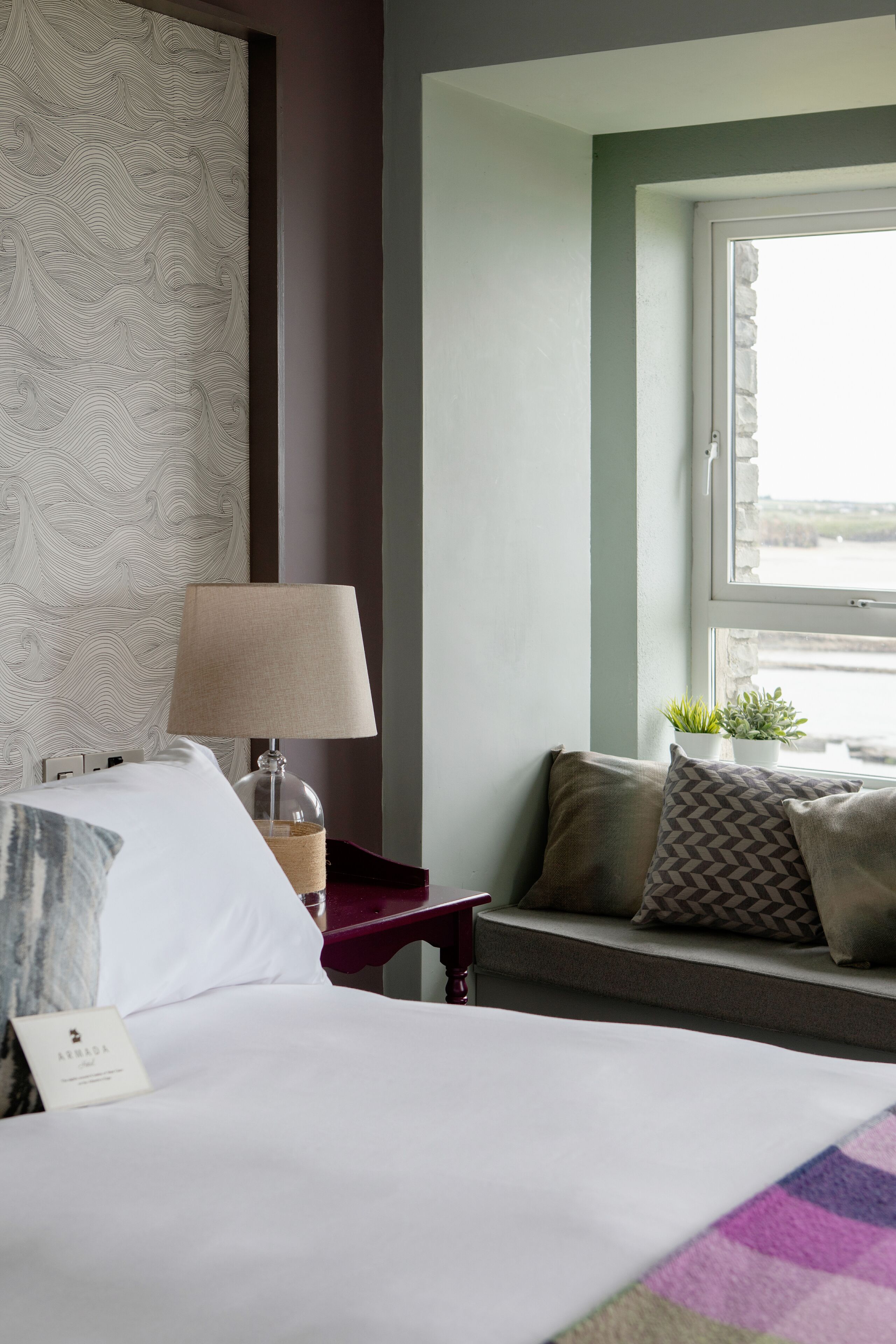 A hotel room with a bed, a nightstand with a lamp, a window with a view, and a sofa with cushions.