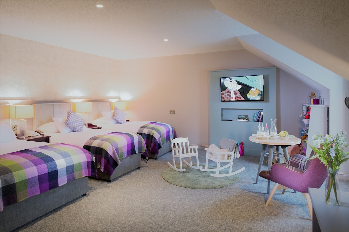 A hotel room with two beds, a TV, a small table with chairs, and a bookshelf.