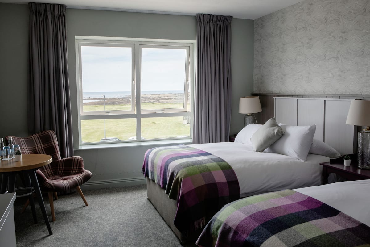A hotel room with a large window, a bed with a colorful blanket, a chair, and a table with a lamp.