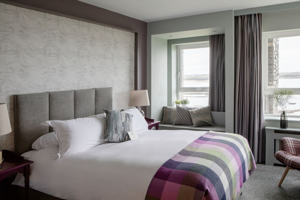 A hotel room with a large bed, a nightstand, a chair, a window with curtains, and a view of the outside.