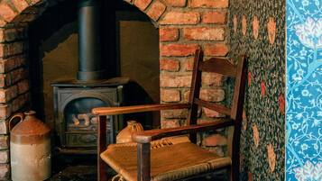 A rustic room with a brick fireplace, a wooden chair, and a shelf with various items including a kettle and a jug.
