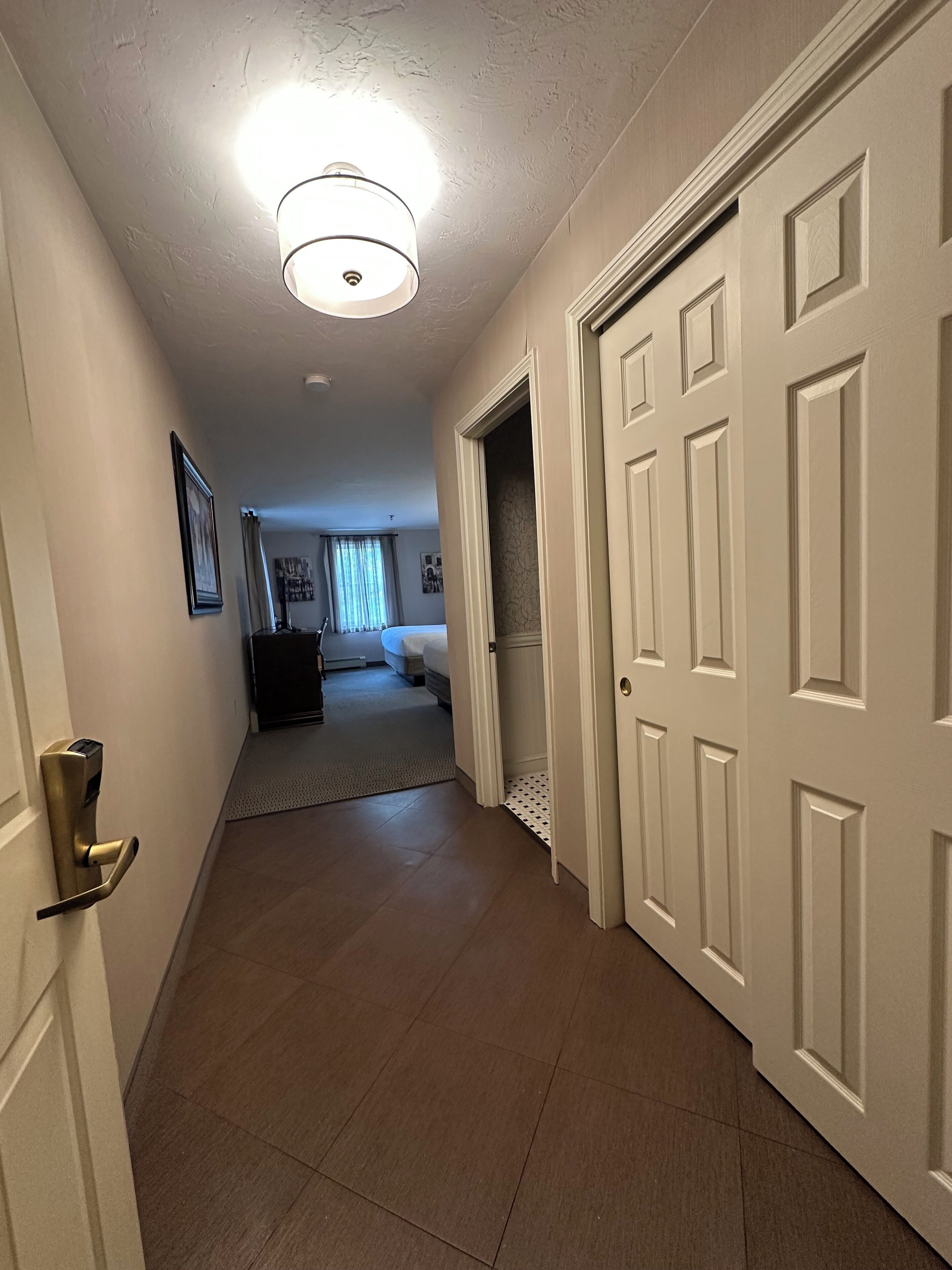 A hallway with a door on the left, a bed in the background, and a ceiling light.
