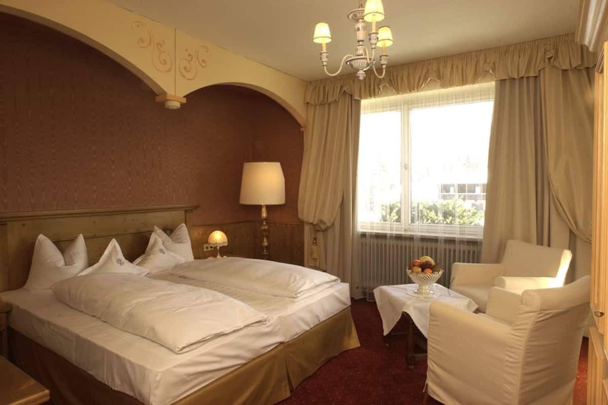 Habitación de hotel con dos camas, un área de estar con una mesa y una silla, un candelabro y una ventana con cortinas.