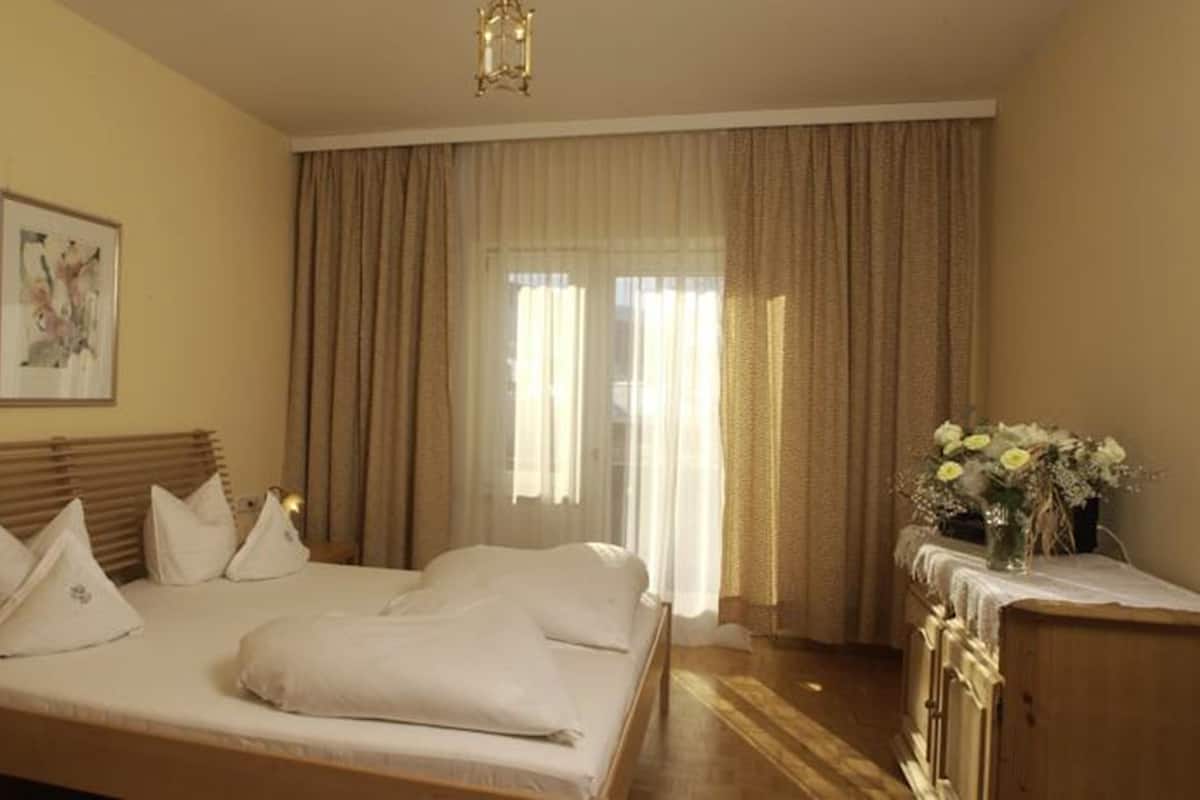 Habitación de hotel con cama, mesita de noche, escritorio, silla, ventana con cortinas y un cuadro en la pared.