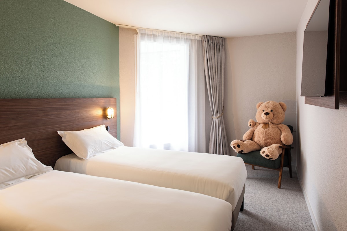 A hotel room with two beds, a large teddy bear on a chair, and a view of the outdoors through a window.