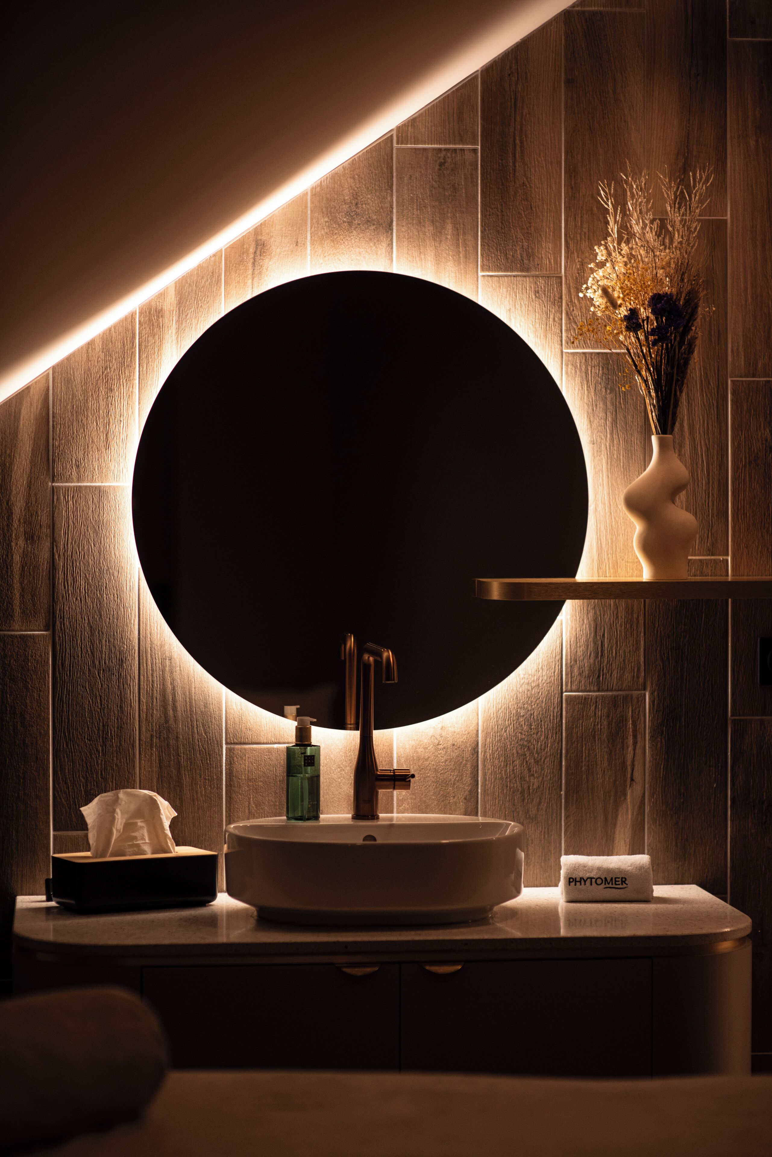 A modern bathroom with a round mirror, wooden wall paneling, a white sink, and a vase with dried flowers.