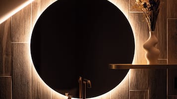 Un baño moderno con un espejo redondo, paneles de madera en la pared, un lavabo blanco y un florero con flores secas.
