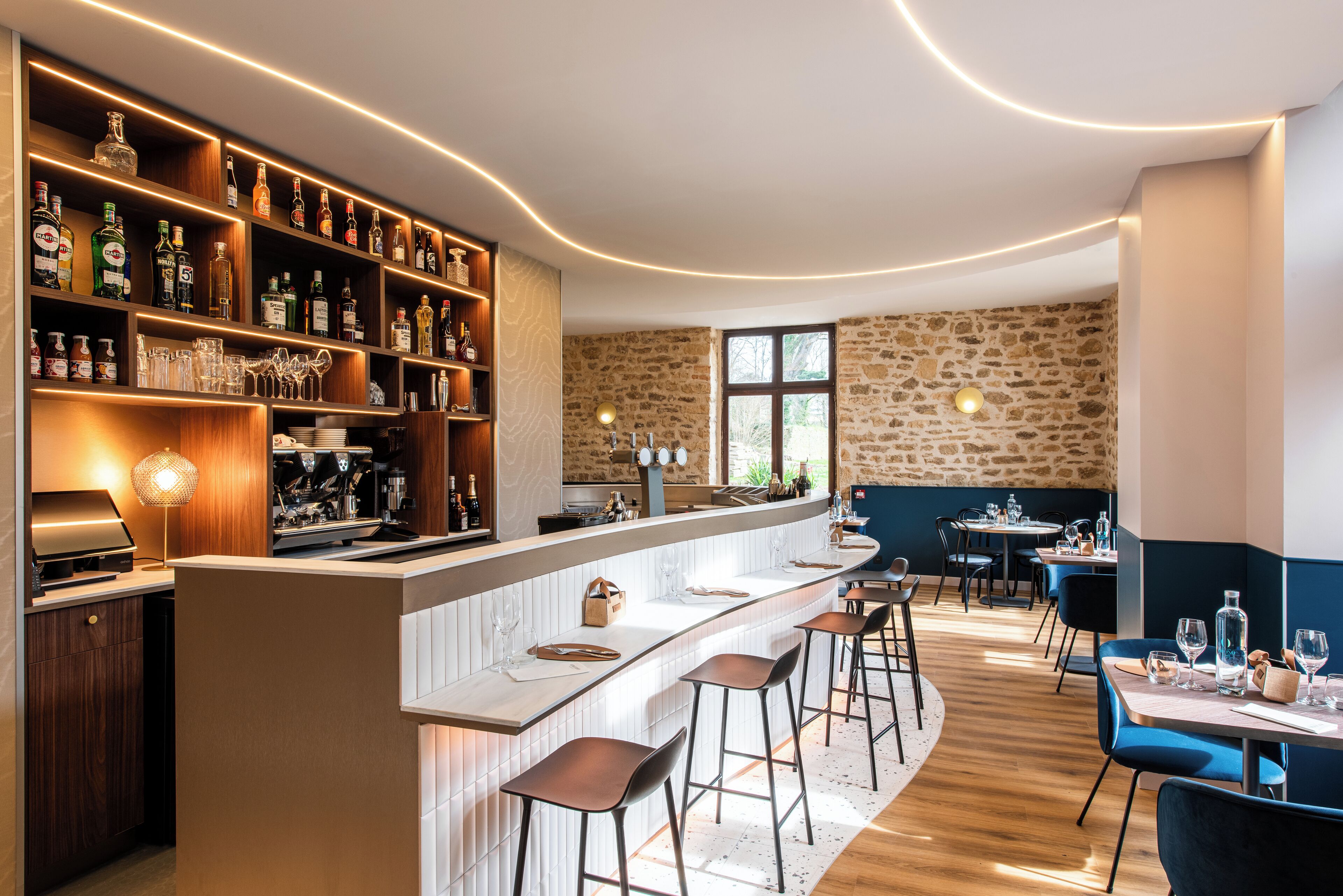 A bar area with shelves of liquor bottles, a counter with stools, and a stone wall in the background.