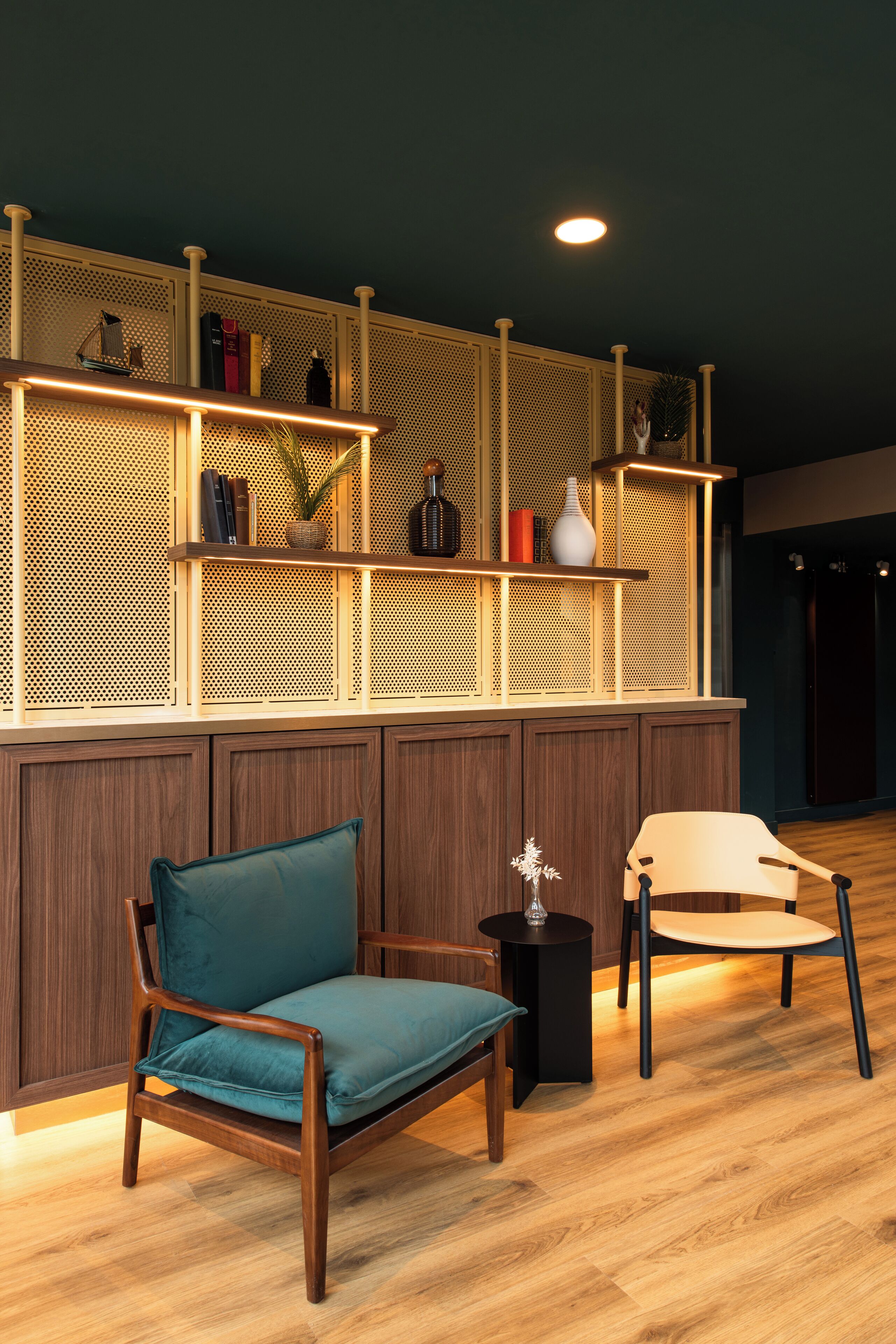 A room with wooden flooring, two chairs, a small table, and a wall-mounted shelf with various items.
