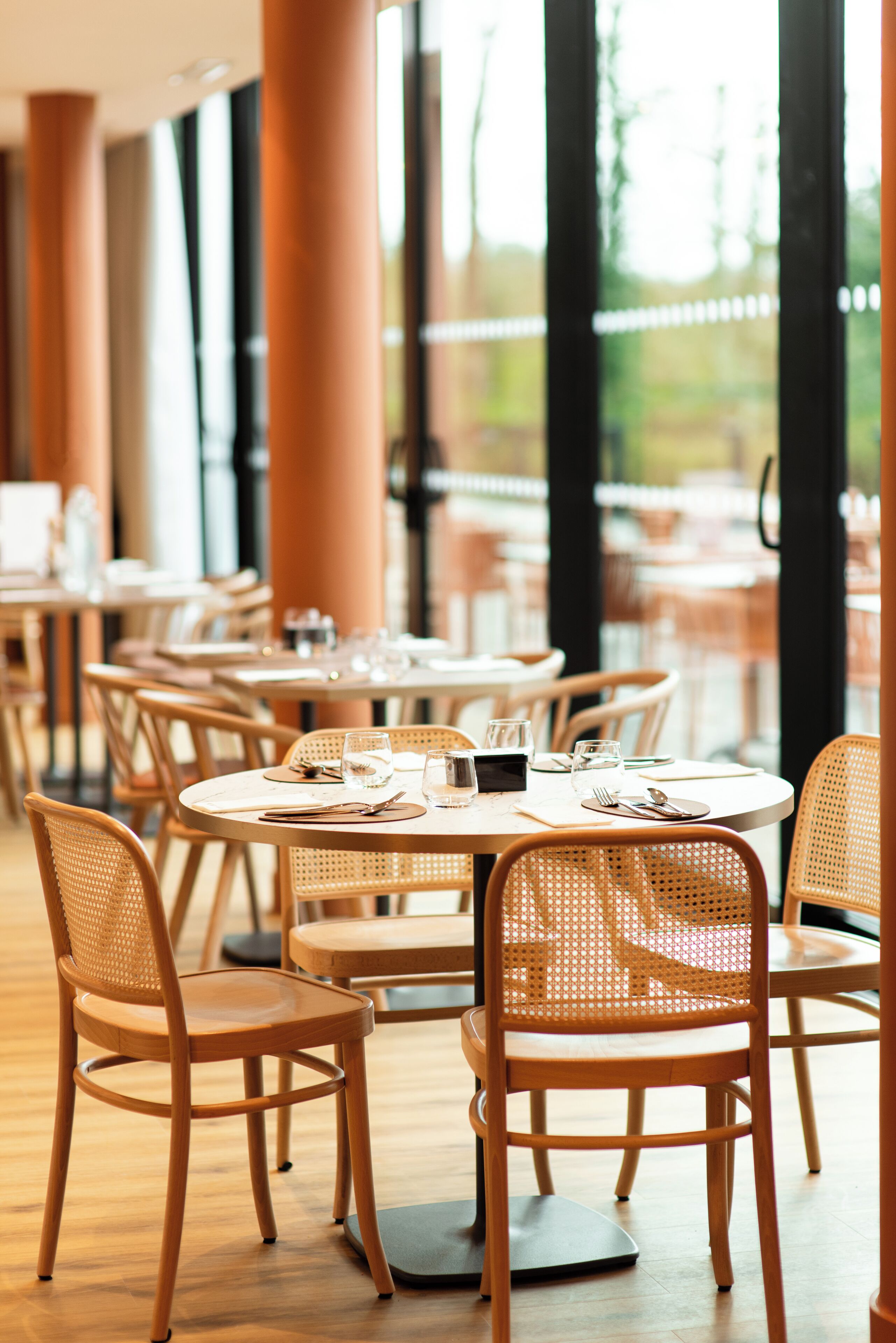 Un restaurante con mesas redondas y sillas de mimbre, ventanas amplias y vistas a zonas verdes.