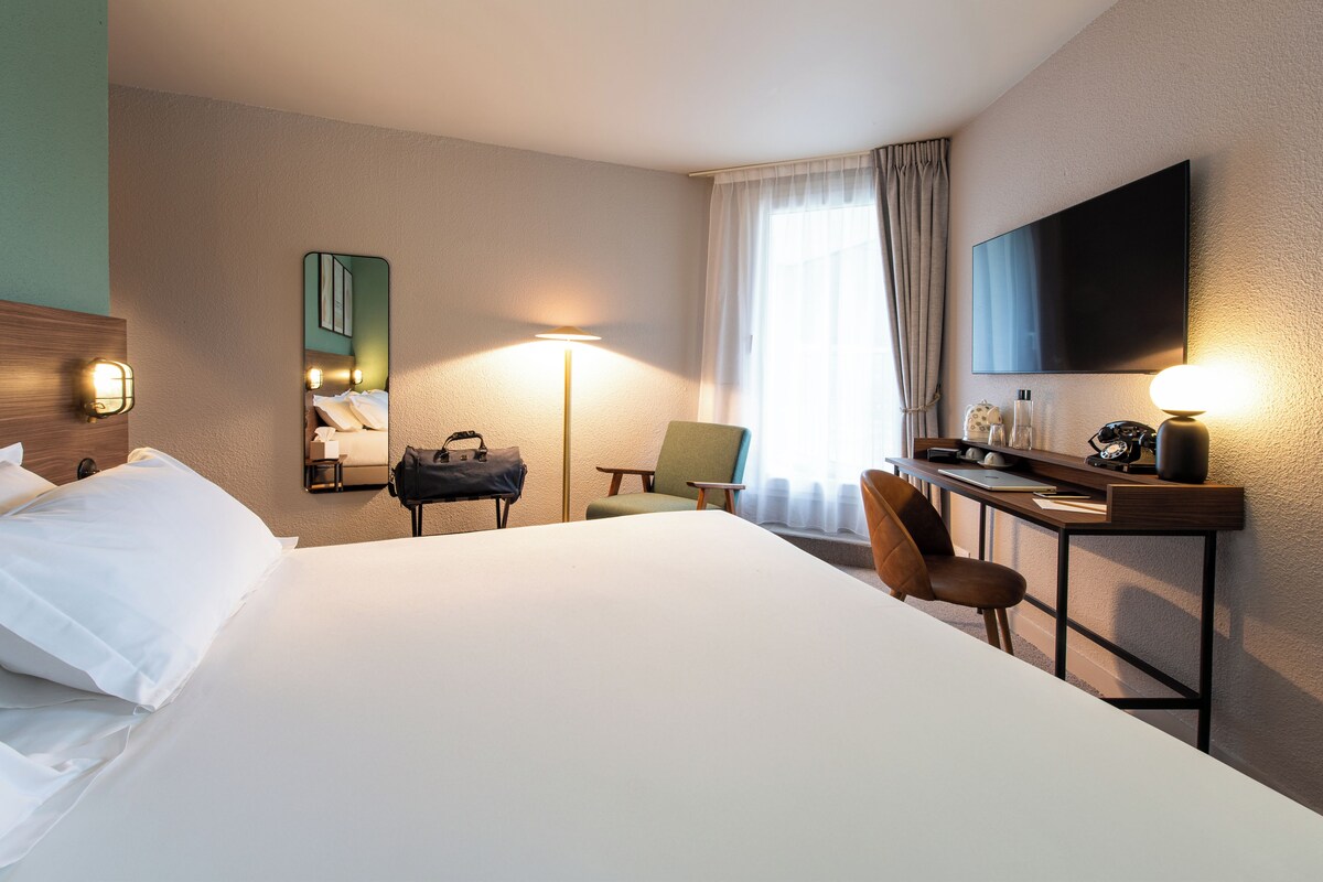Une chambre d’hôtel avec un grand lit, un bureau avec une chaise, un téléviseur à écran plat, une lampe et un miroir.