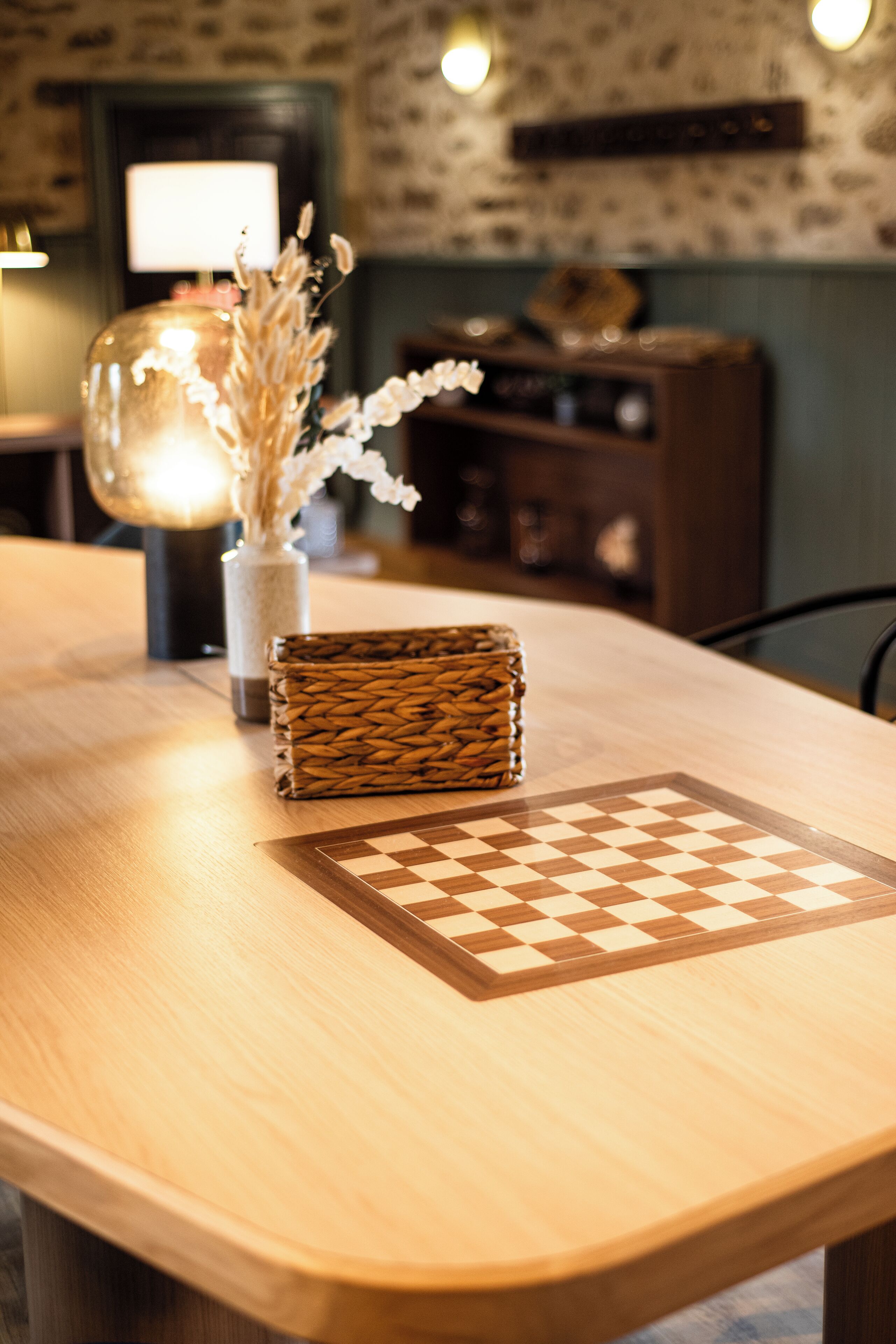 A wooden table with a chessboard, a woven basket, and a vase with dried flowers.