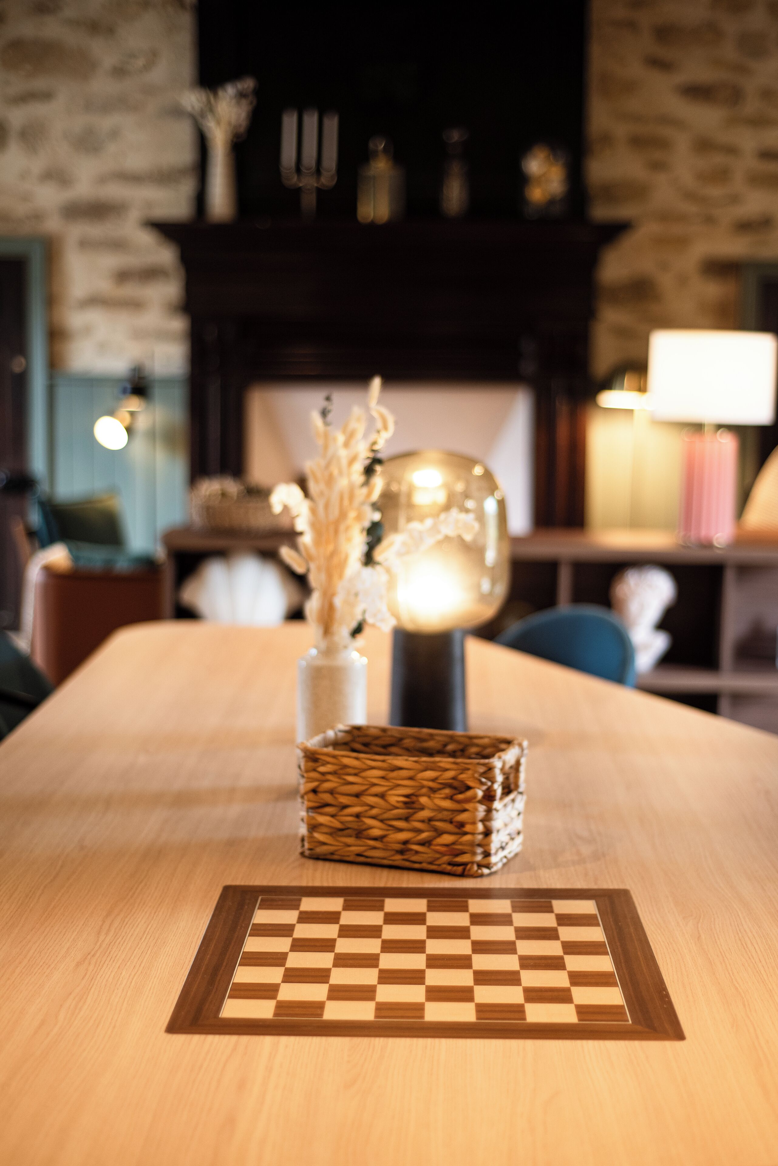 Una mesa de madera con un tablero de ajedrez, un florero con plantas secas y una lámpara esférica.