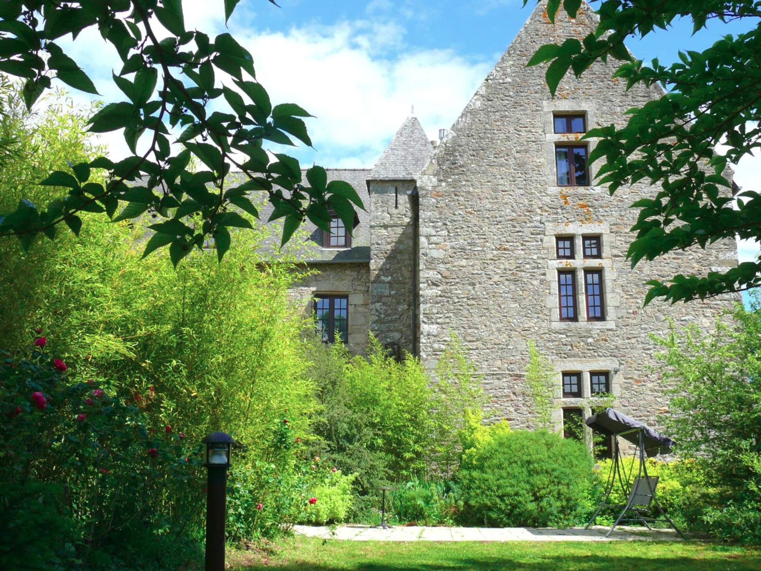 A stone building with multiple windows and a garden with lush greenery and red flowers.
